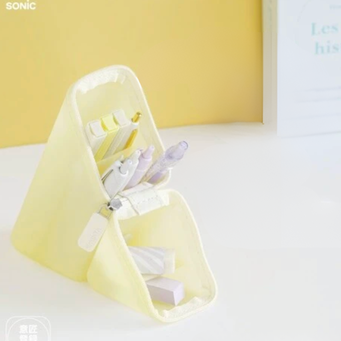 A pale yellow fabric desk organizer with multiple compartments holding pens and stationery, set on a white surface against a soft yellow background.