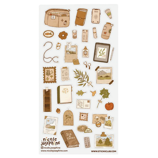 A sheet of Nicole Josephine 'Six Months' travel-themed sticker designs, featuring illustrations of a suitcase, journal, camera, map, leaves, and an apple in a warm, vintage brown and beige palette on a white background.
