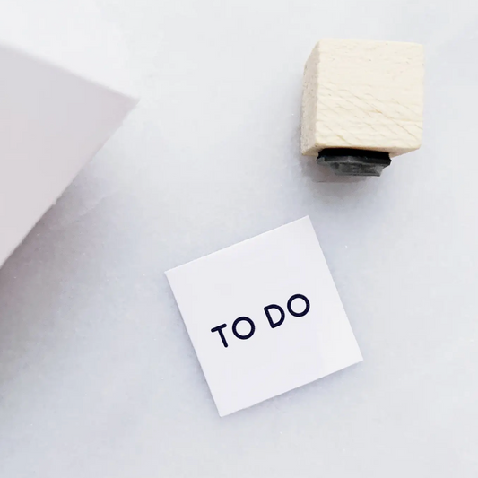 A small white sticky note with the words 'TO DO' printed in black, placed on a light gray surface next to a cream-colored wooden desk stamp.