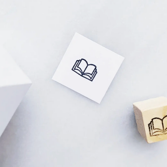 A small white square card printed with a simple black outline of an open book, placed on a light gray surface. A wooden stamp block with a matching open-book design is partially visible at the lower right.