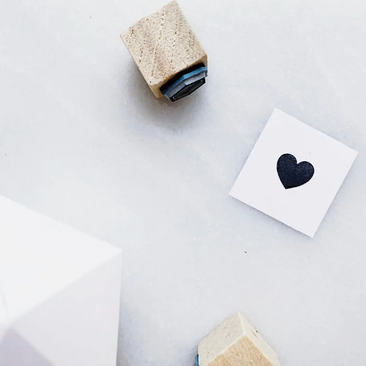 A styled flat-lay of a small white square card featuring a black heart, alongside two wooden cube stamps with blue and beige inks, on a light gray background.