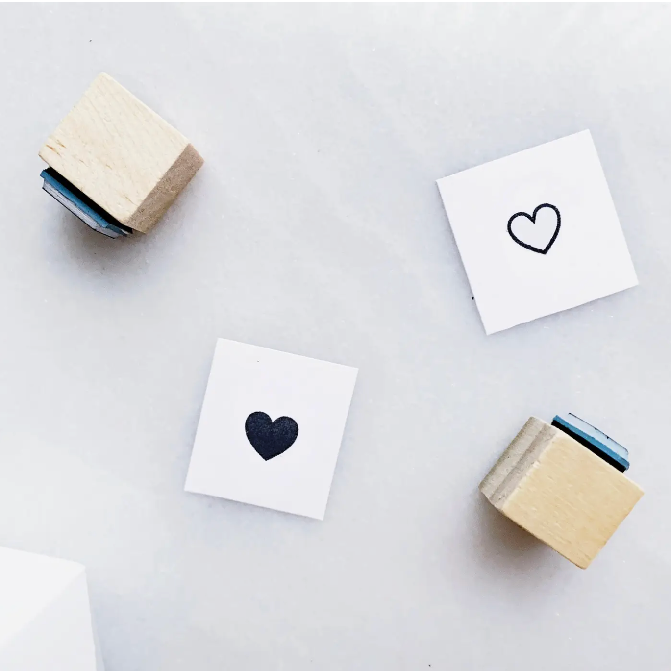 Two wooden rubber stamp blocks with blue handles are arranged on a light gray surface, with two small white cards showing black heart stamp impressions.