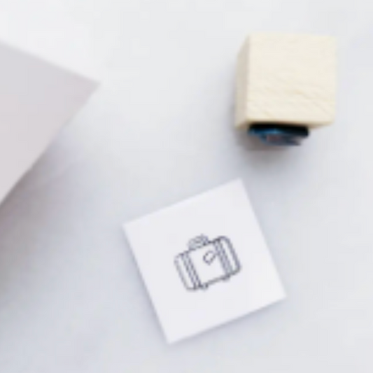 A small white square card printed with a simple black suitcase outline, placed beside a wooden-handled stamp and a folded white sheet on a light gray surface.