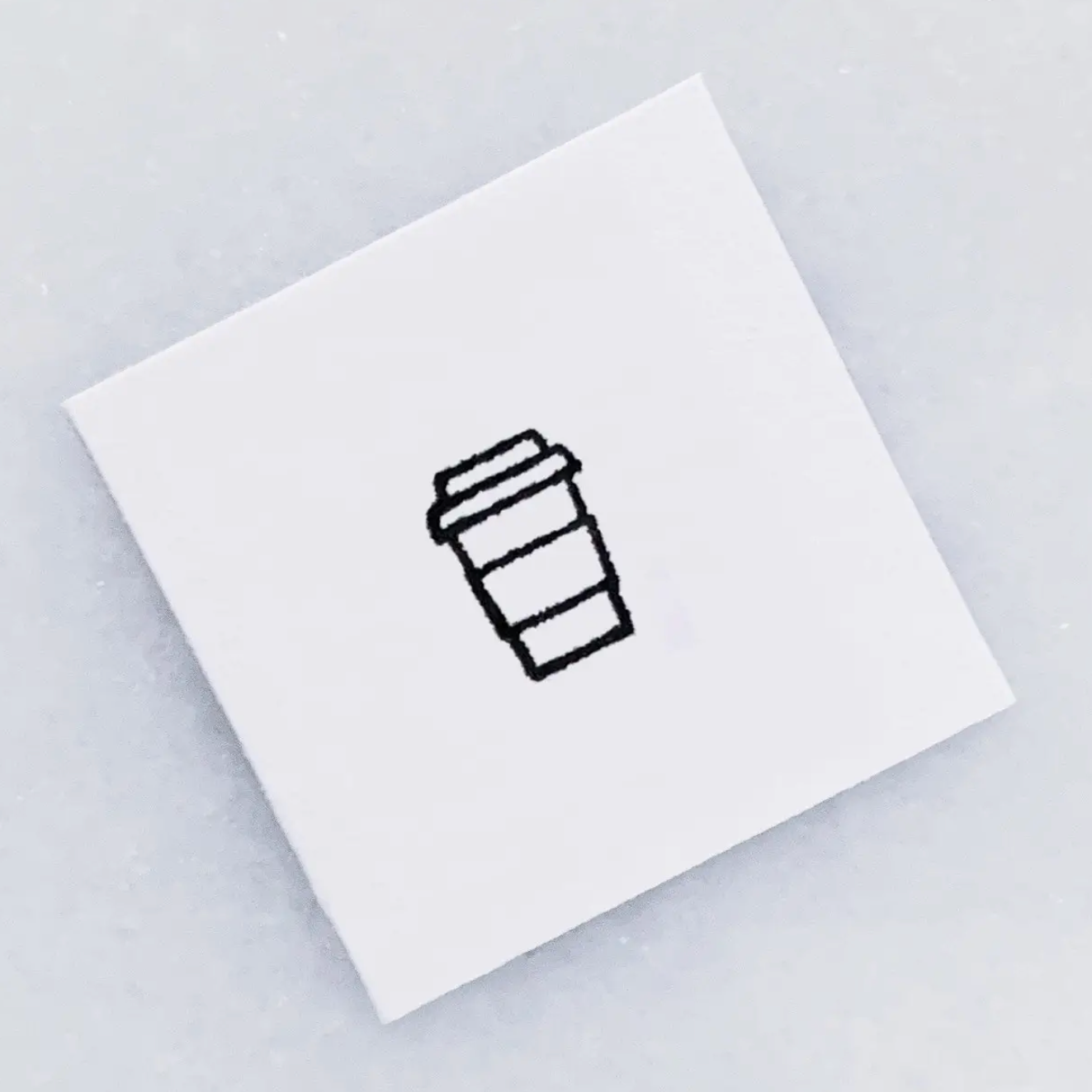 The top of a  white square stamp with a simple black line drawing of a takeaway coffee cup, photographed on a light gray background.