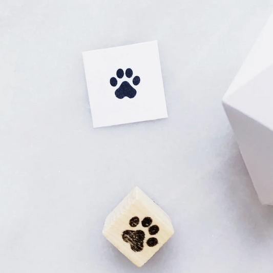 A small square cream-colored item with a dark paw print mark, shown on a light gray surface next to a white square card with a matching paw print and part of a white box.