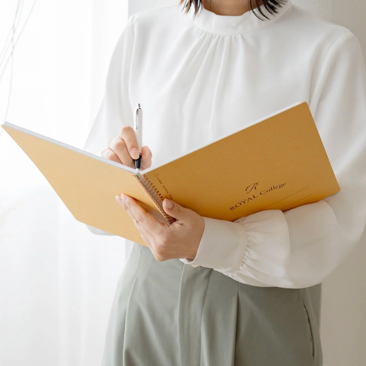 A person in a white blouse and light gray skirt holds an open mustard-yellow notebook with a pen, standing near a bright window.