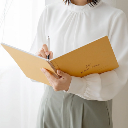 A person in a white blouse and light gray skirt holds an open mustard-yellow notebook with a pen, standing near a bright window.