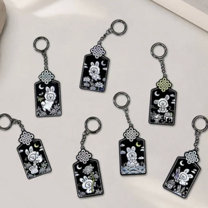 Six black enamel keychains featuring white line-art illustrations of rabbits, crescent moons, and flowers, arranged on a light beige surface.