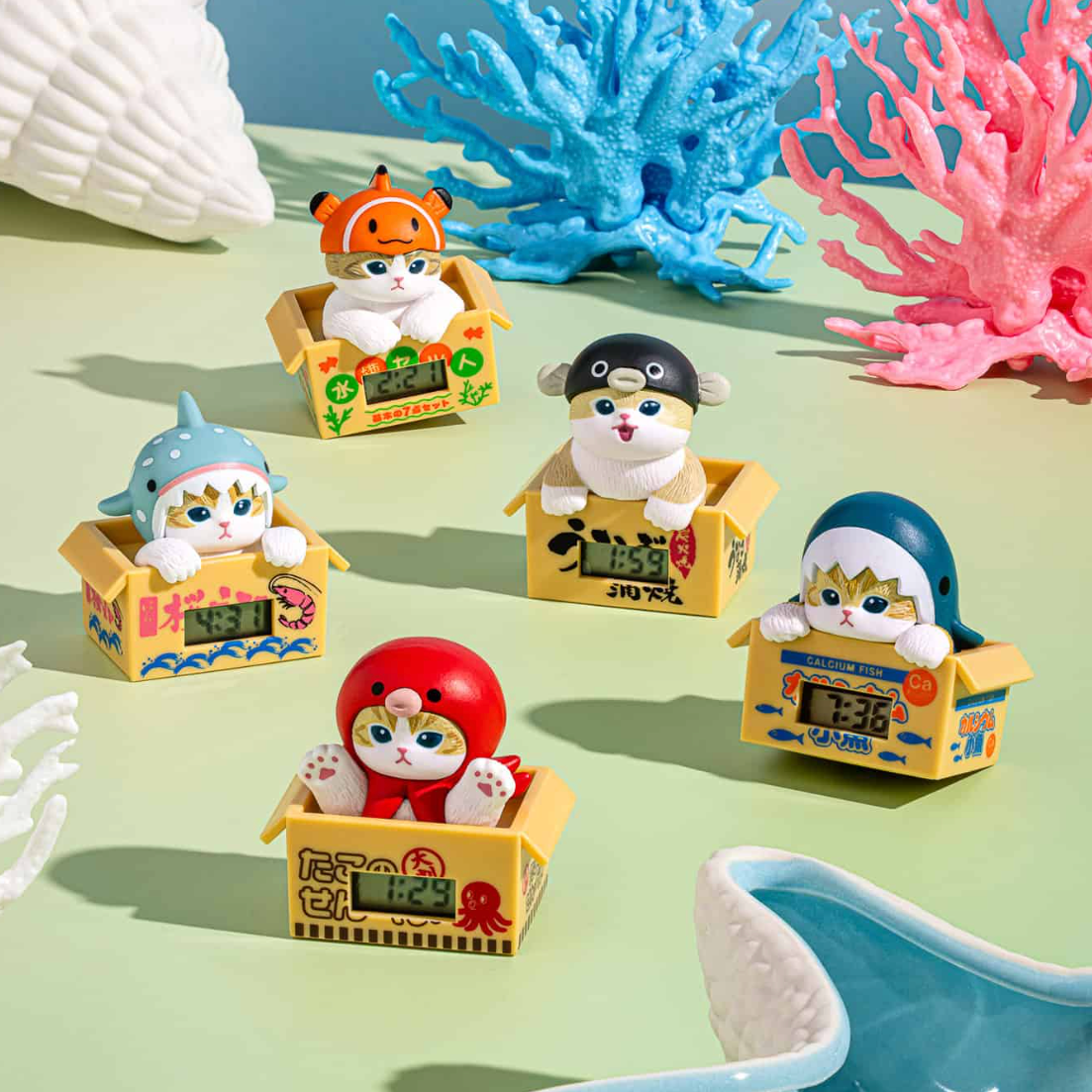 Six small cat figurines dressed as sea creatures sitting in yellow boxes, arranged on a mint green surface with coral and shell props.