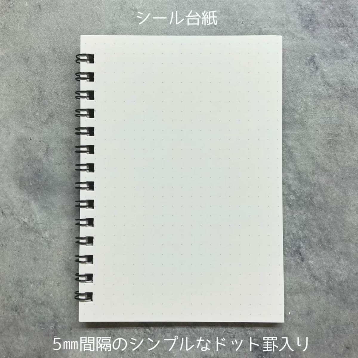 A white dotted notebook sheet with a black spiral binding, shown flat on a gray textured background with Japanese text at the top and bottom.