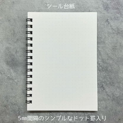 A white dotted notebook sheet with a black spiral binding, shown flat on a gray textured background with Japanese text at the top and bottom.