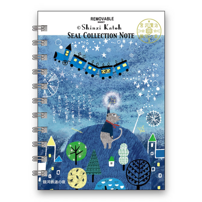 A spiral-bound notebook titled 'REMOVABLE Seal Collection Note' by Shinzi Katoh, featuring a whimsical blue winter illustration with a train, cat, trees, and stars on the cover.