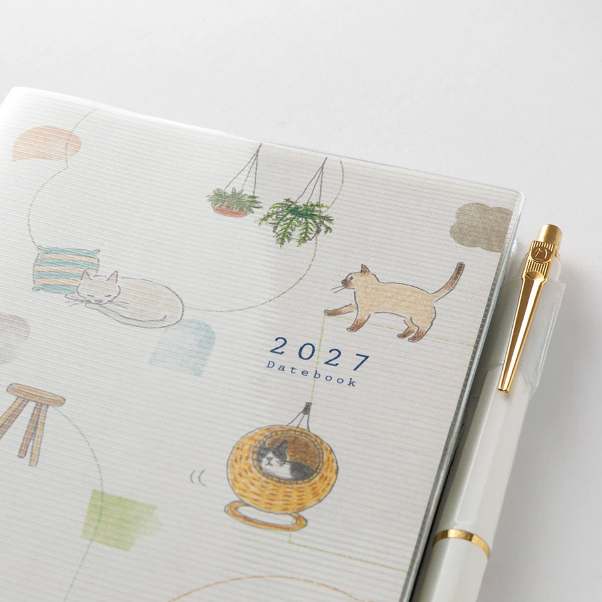 A 2027 datebook with a white ribbed cover decorated with cute cat illustrations and small hanging plant drawings, shown next to a white pen with gold accents on a clean white background.