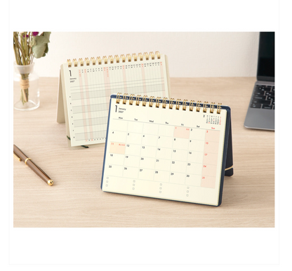 A spiral-bound desk calendar shown open on a light wood tabletop, with a second calendar standing behind it. The front calendar has a navy blue cover and an off-white page, with a few highlighted dates in soft pink.