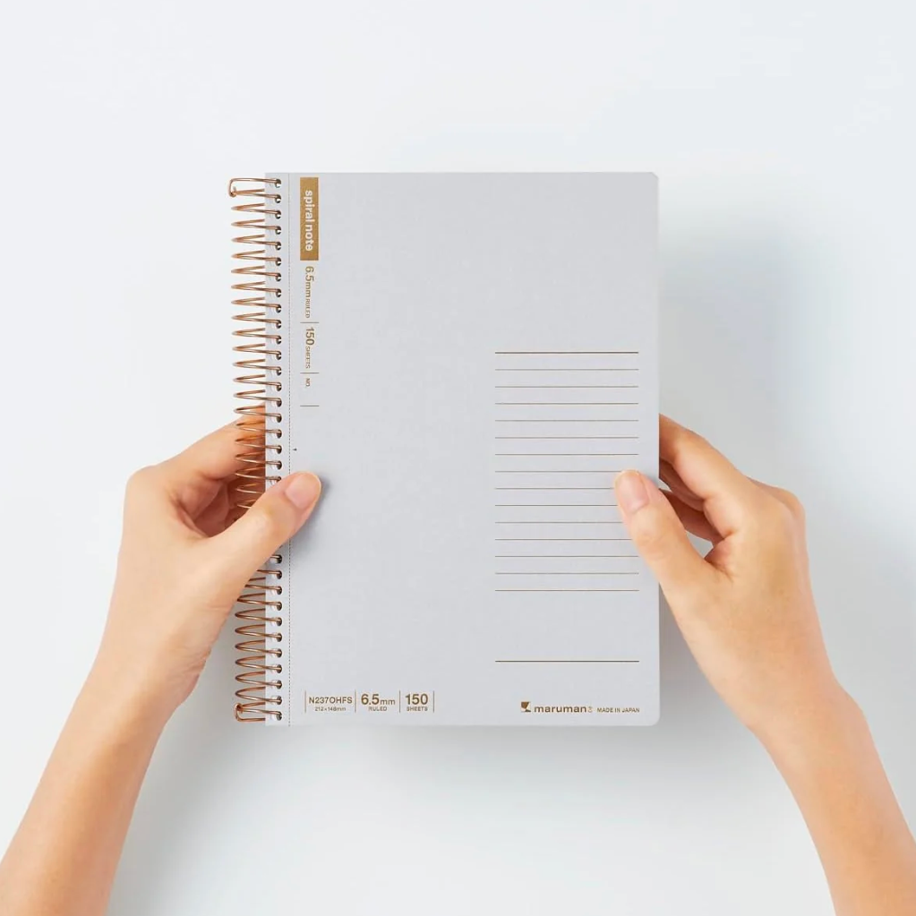 A person holds a light gray spiral-bound planner against a clean white background. The planner has a rose gold wire coil binding, gold accents, and a lined page layout on the cover.