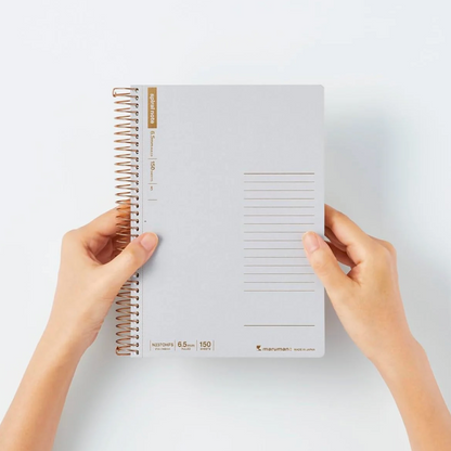 A person holds a light gray spiral-bound planner against a clean white background. The planner has a rose gold wire coil binding, gold accents, and a lined page layout on the cover.