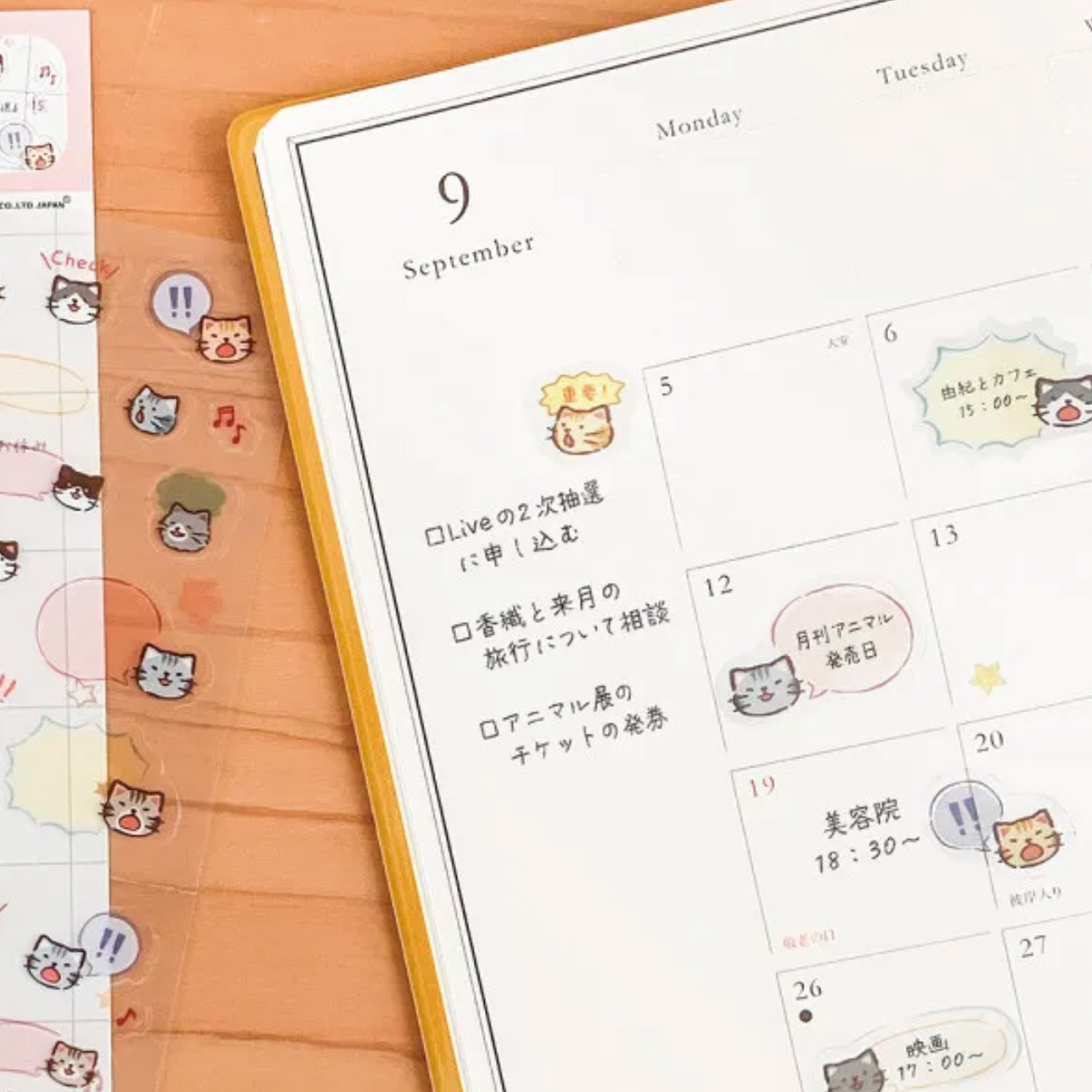 A close-up of a planner or calendar page with cute cartoon cat stickers and handwritten notes, placed on a peach-colored desk next to a sheet of matching sticker strips.