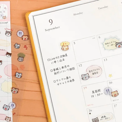 A close-up of a planner or calendar page with cute cartoon cat stickers and handwritten notes, placed on a peach-colored desk next to a sheet of matching sticker strips.