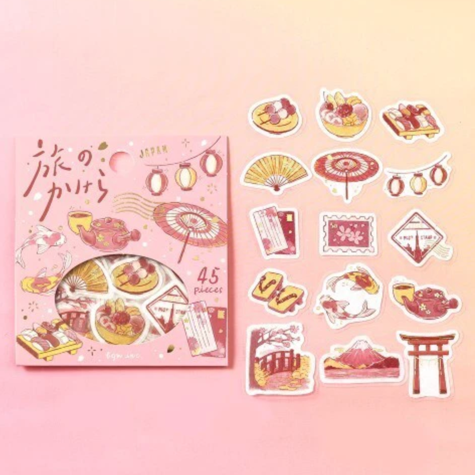 A pink sticker set package labeled 'JAPAN' with a circular window showing assorted travel-themed stickers, placed beside the package on a soft pink background. The stickers depict Japanese motifs including lanterns, umbrellas, food, postcards, mountains, and torii gates in pink, red, and beige tones.