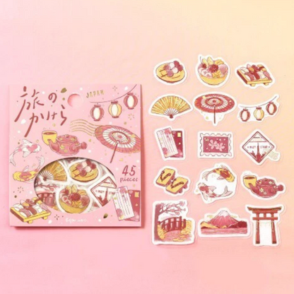 A pink sticker set package labeled 'JAPAN' with a circular window showing assorted travel-themed stickers, placed beside the package on a soft pink background. The stickers depict Japanese motifs including lanterns, umbrellas, food, postcards, mountains, and torii gates in pink, red, and beige tones.