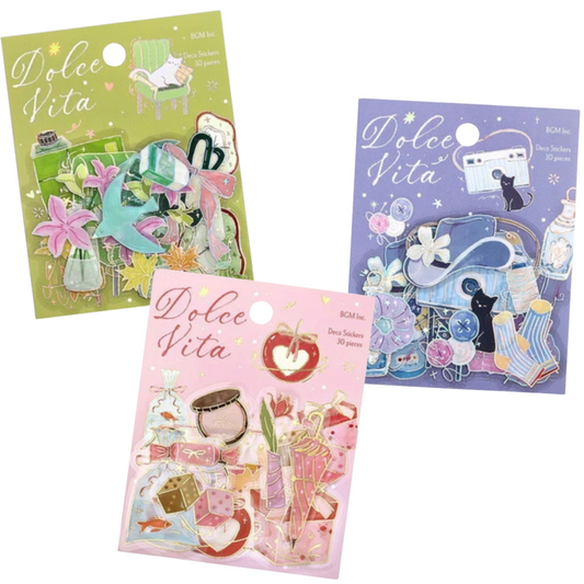 Three packaged sticker sheets from Dolce Vita, each with a different pastel background color: olive green, lavender, and pink. The sheets feature whimsical illustrated stickers including flowers, hearts, stars, and small objects, and are shown on a plain white background.