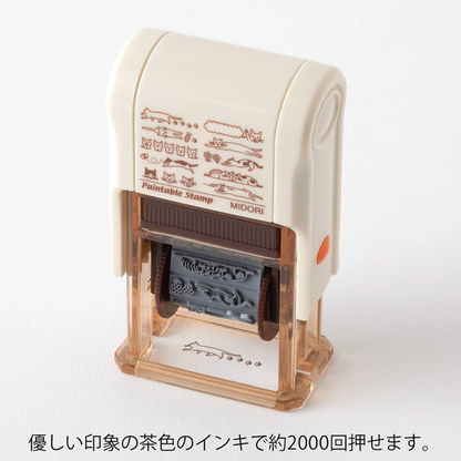 A tabletop ink stamp with a cream-colored plastic body, clear transparent base, and brown printed sample graphics on the front. Japanese text appears below the product on a white background.