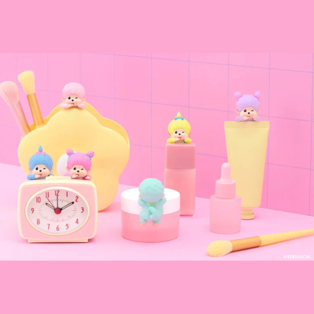 A pastel pink and yellow cosmetic-themed miniature set featuring small character figurines on top of makeup items, including a yellow makeup sponge holder, a pink alarm clock, a pink bottle, a dropper bottle, a cream jar, and a brush, arranged on a pink surface against a pink tiled background.
