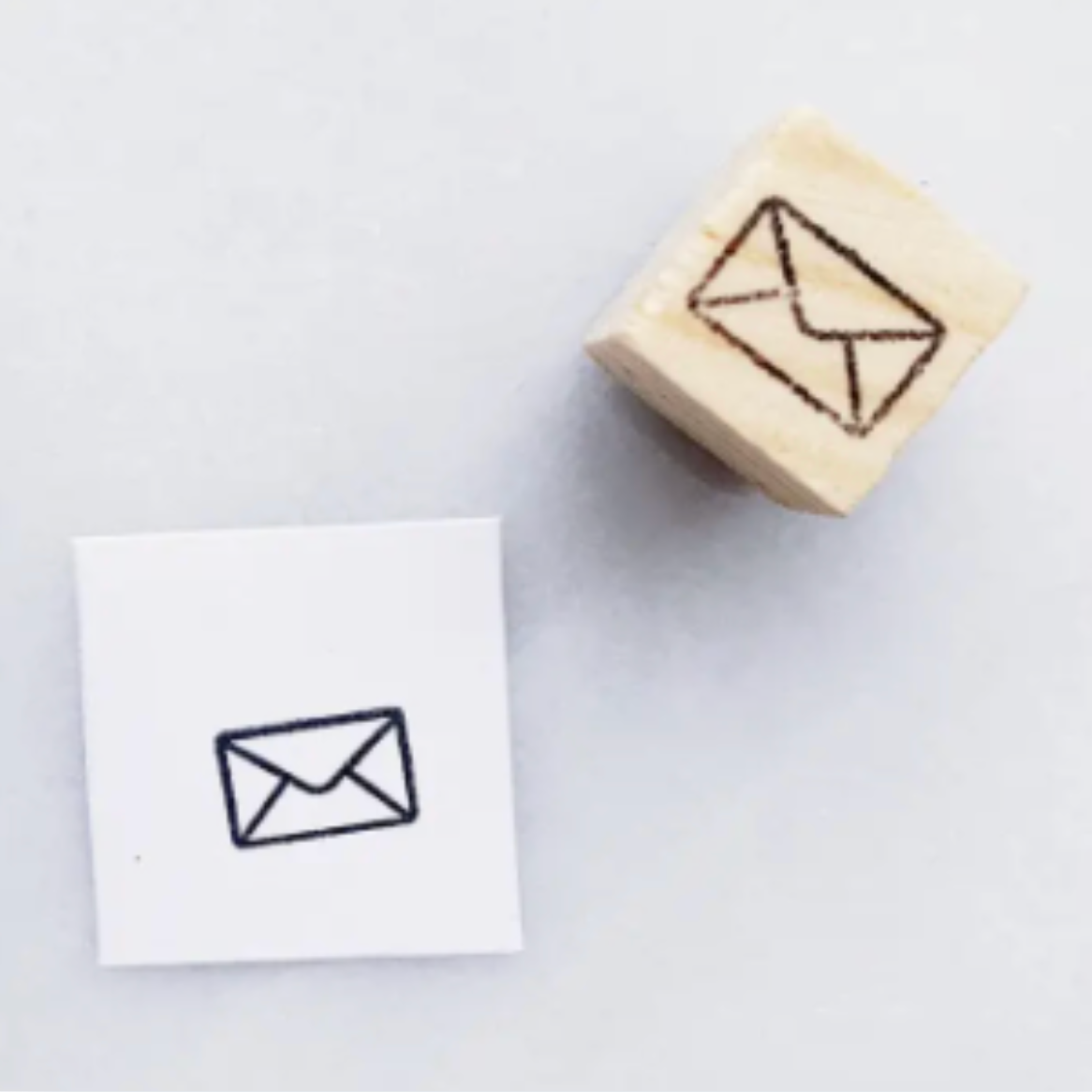 A wooden rubber stamp with an envelope icon and a small white card showing the stamped black envelope mark, photographed on a light gray background.