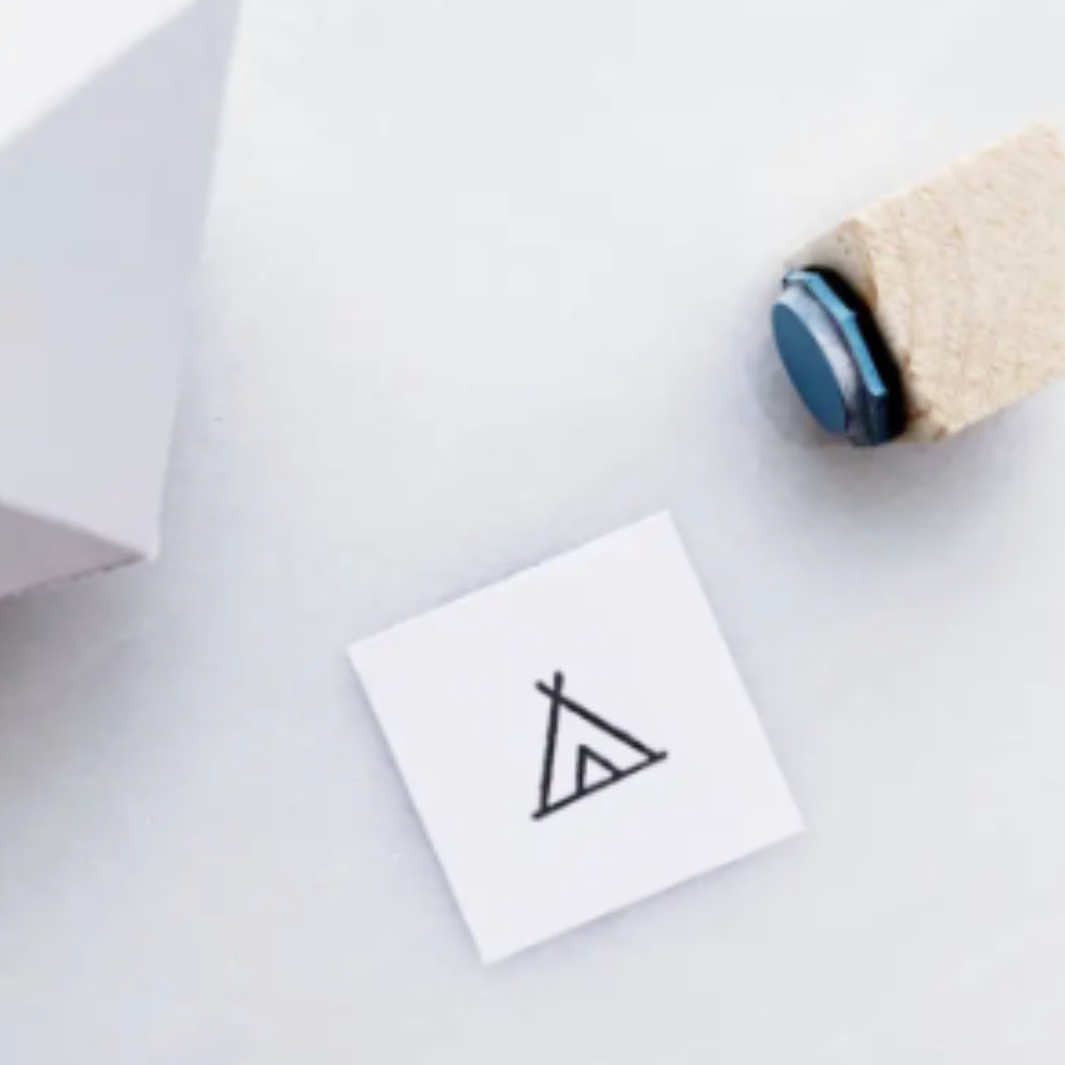 A small white square card printed with a simple black tent-like symbol, placed on a light gray surface next to a white box corner and a rolled natural-fiber pen or stamp handle with a blue circular end.