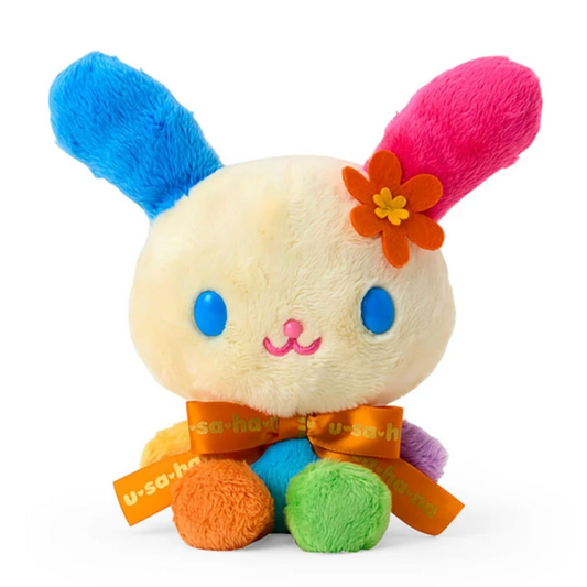 A plush bunny toy with cream-colored fur, bright blue and pink ears, a small orange flower on the head, blue eyes, and colorful pom-pom feet. It wears an orange ribbon with 'usabii' text, set against a plain white background.