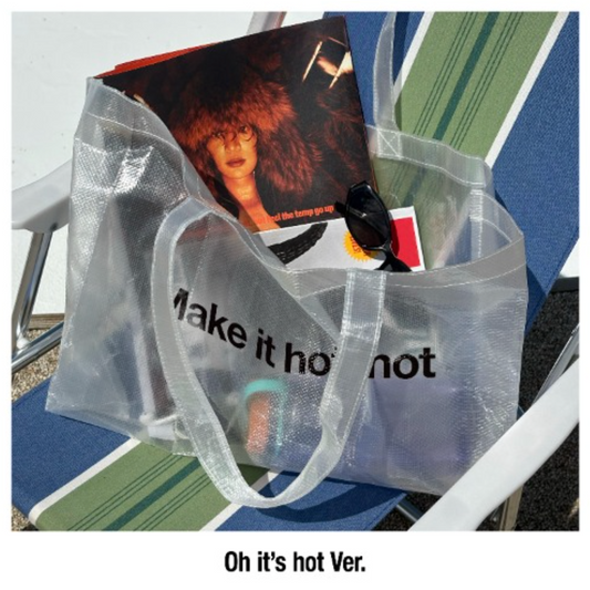 A translucent clear tote bag with black text reading 'Make it hot not' sits on a blue and green striped lounge chair outdoors. A magazine, sunglasses, and other items are tucked inside the bag.