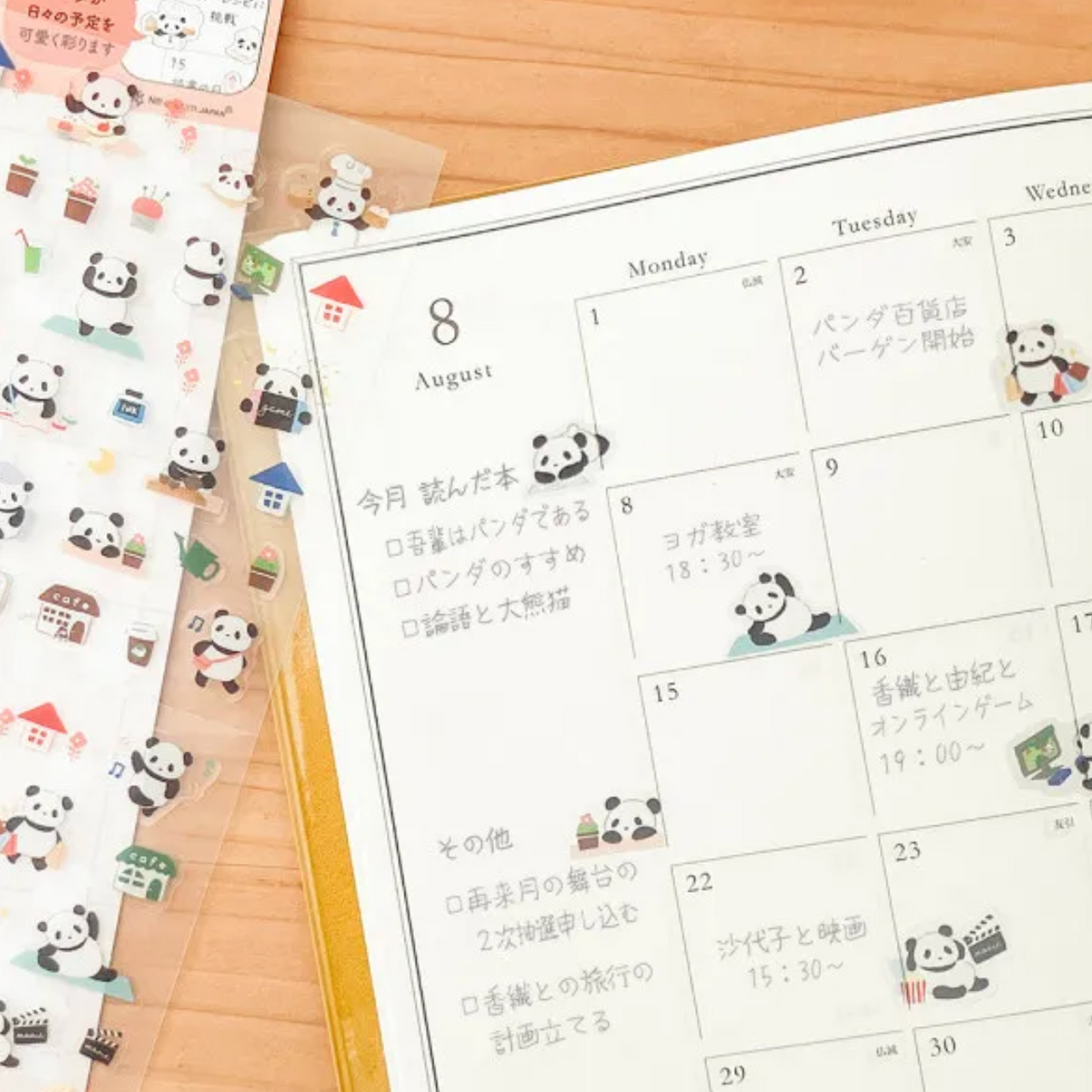 A close-up of a Japanese planner or calendar page with panda-themed stickers applied to dates and notes, placed on a wooden surface next to a sheet of unapplied stickers.