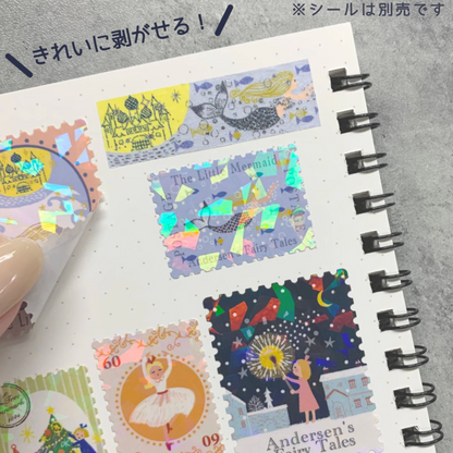 A close-up of a spiral notebook page with several colorful postage-stamp-style stickers featuring whimsical fairy tale illustrations, including a mermaid, ballerina, and starry night scene. A hand is peeling back one sticker, and Japanese text appears at the top.