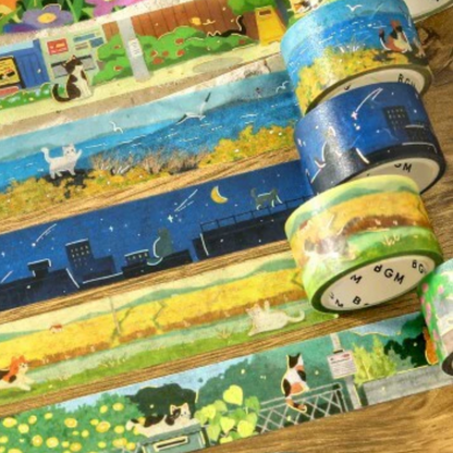 A collection of decorative washi tapes with whimsical cat-themed illustrations, including outdoor scenes, rooftops, and cozy outdoors, arranged on a wooden surface.